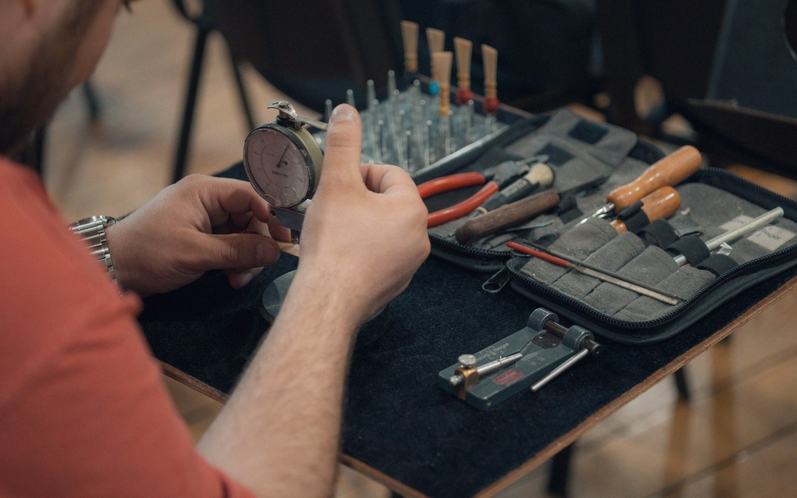 A watchmaker examines a dial gauge beside a tool case filled with pliers and screwdrivers