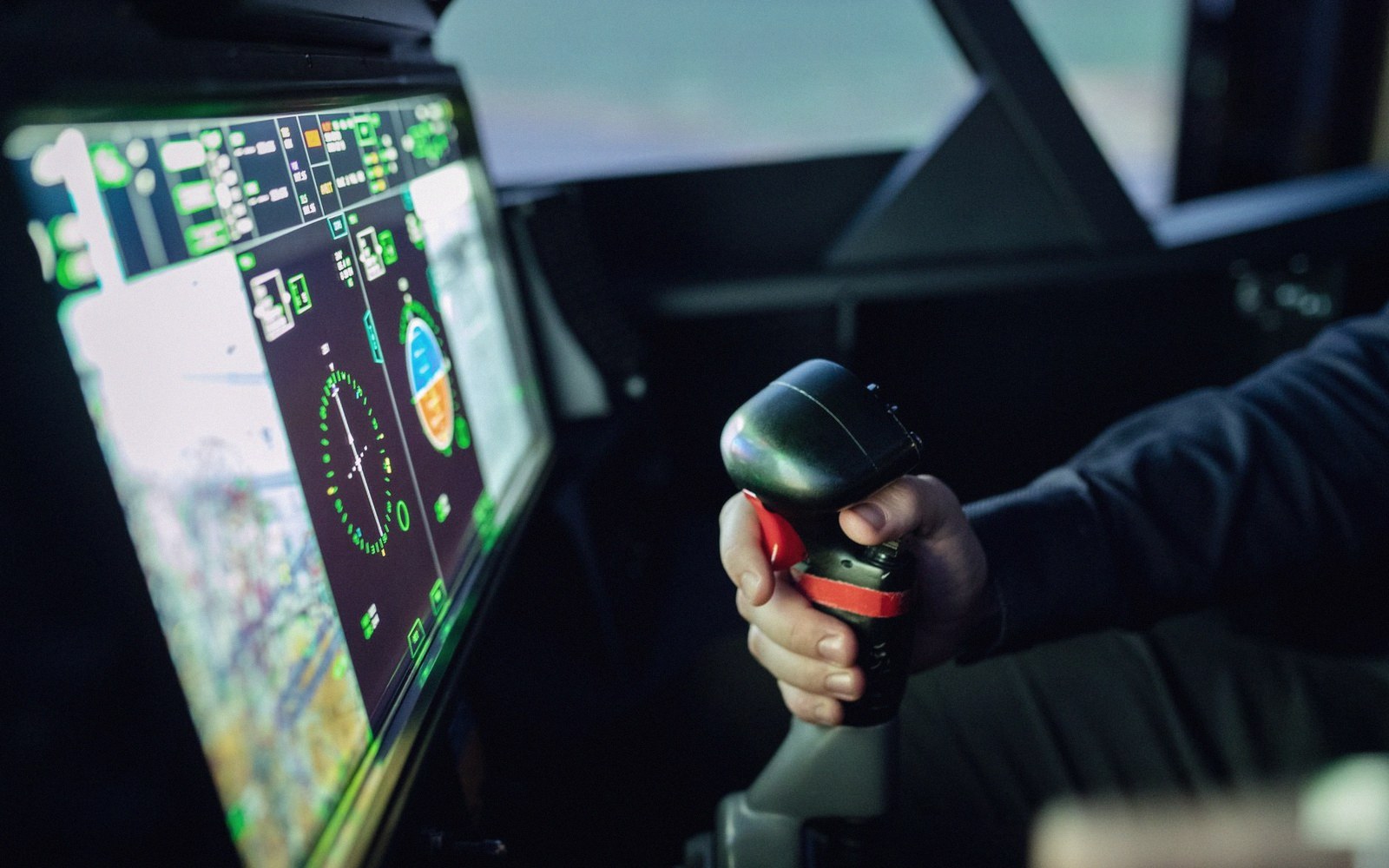 A pilot's hand grips a flight control stick beside a glowing cockpit display